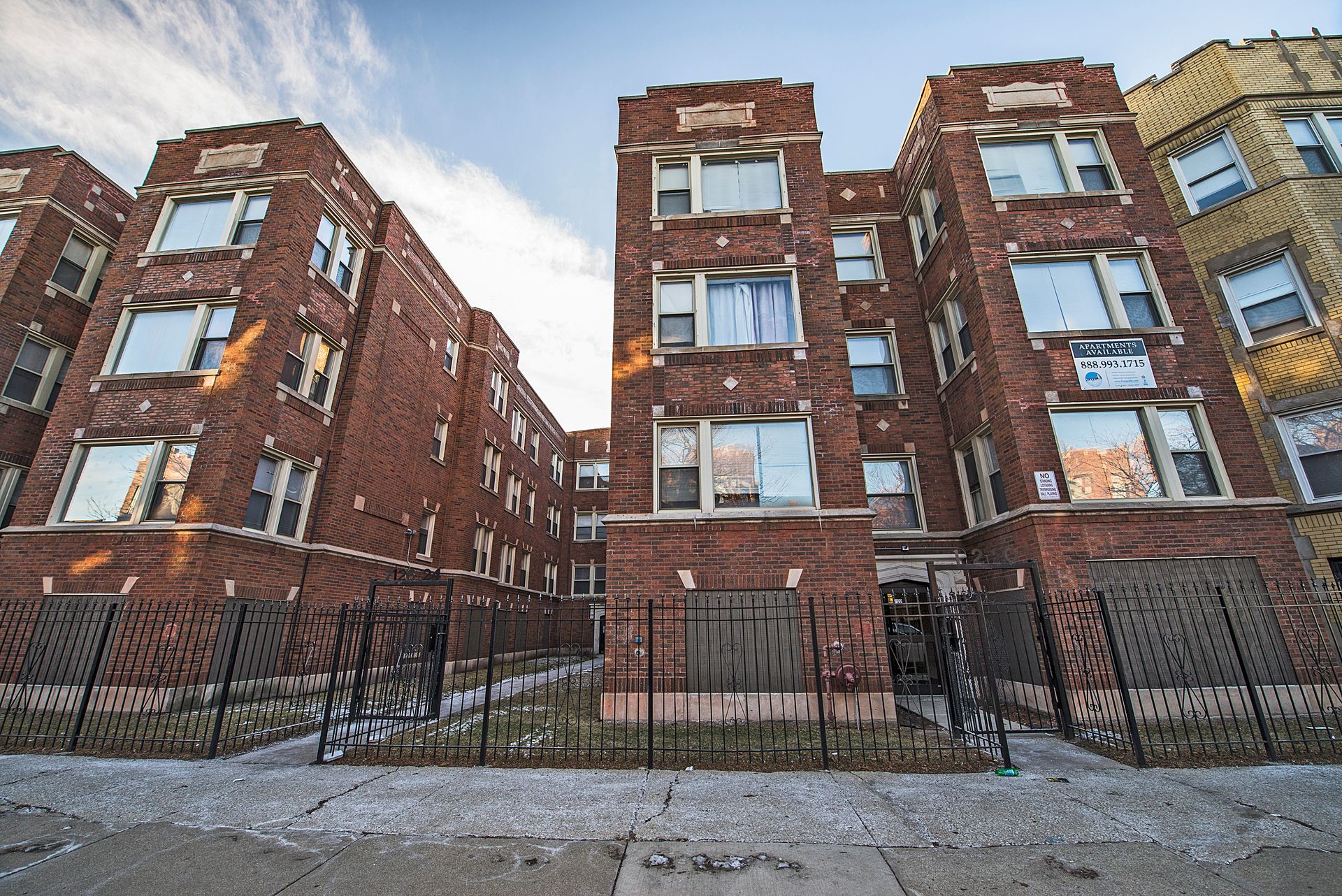 Brick apartment buildings, fenced yard, cloudy sky.