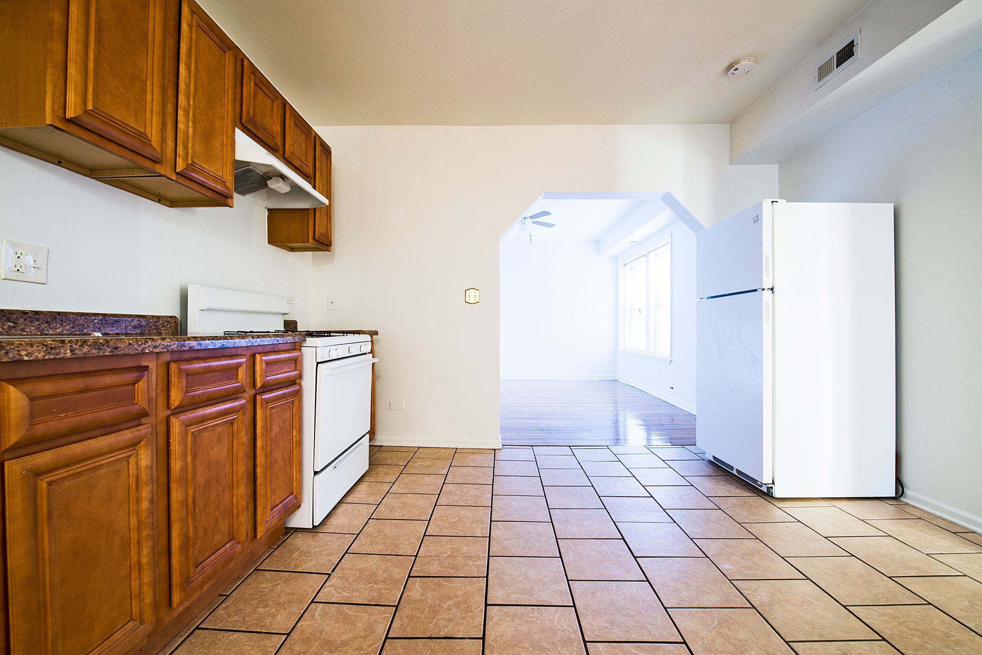 Kitchen with brown cabinets, white appliances, and tan tile floor, leading to a living area.