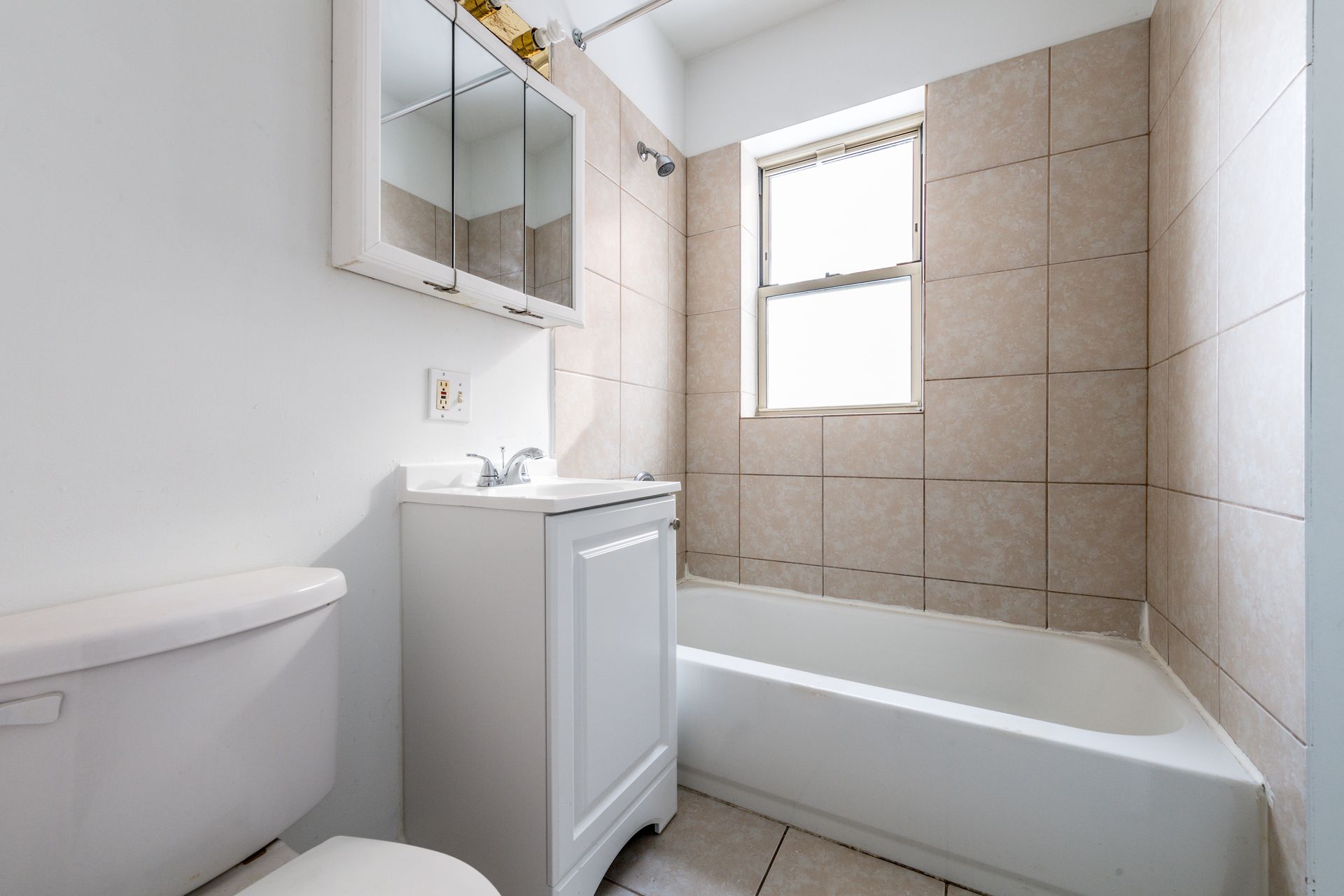 Bathroom with white toilet, vanity, and bathtub. Beige tiled walls. Window on right.