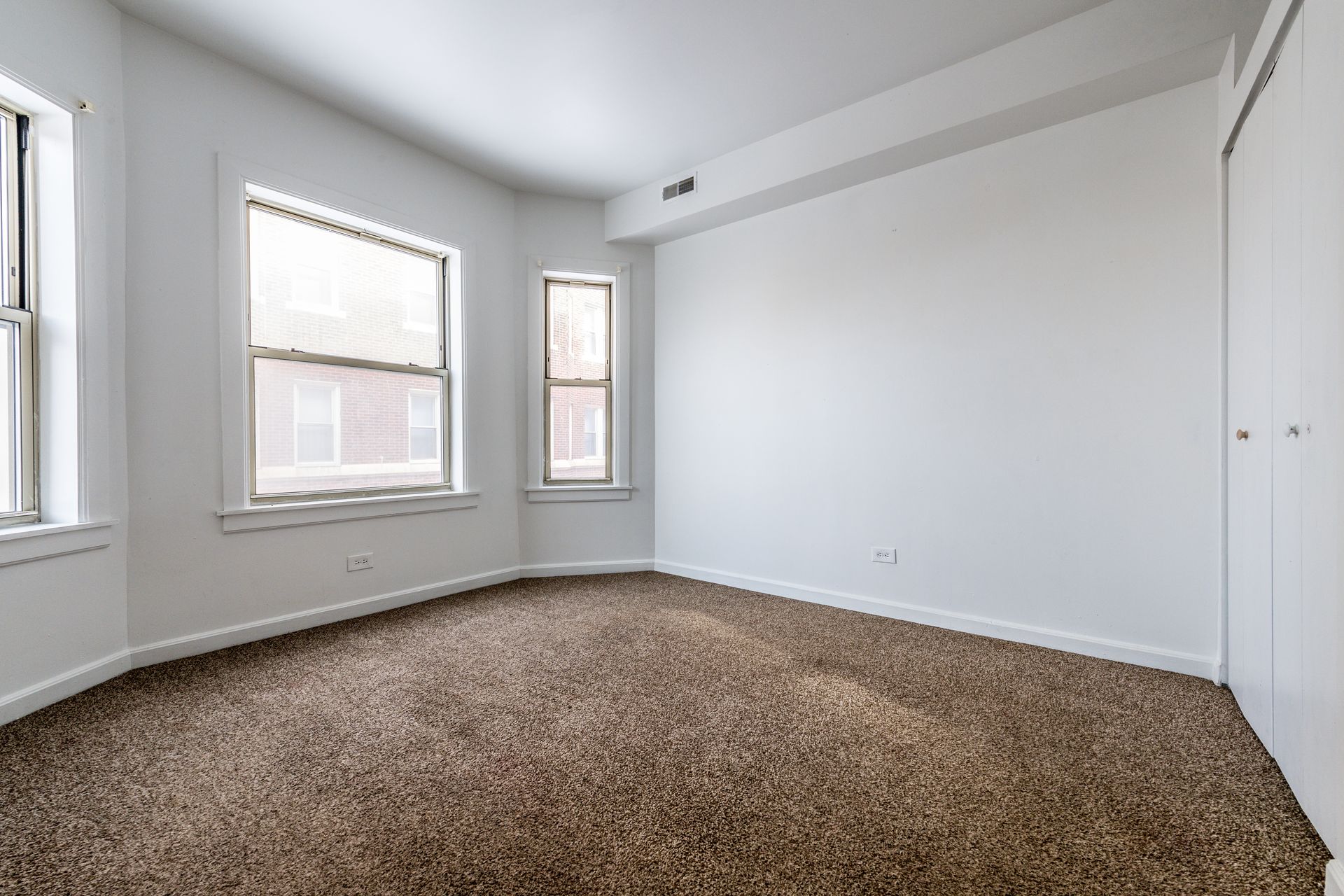 Empty room with brown carpet, white walls, and three windows. A closet is on the right side.