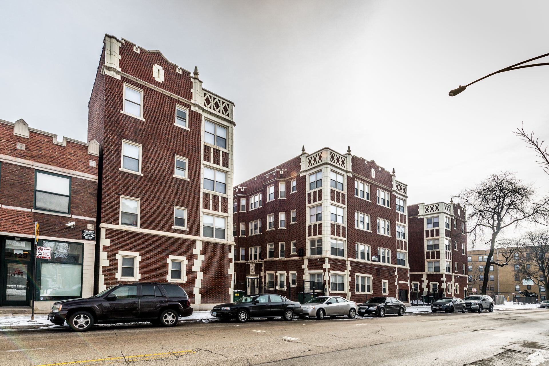 Apartment buildings on a city street, brick facades, cars parked, overcast sky.