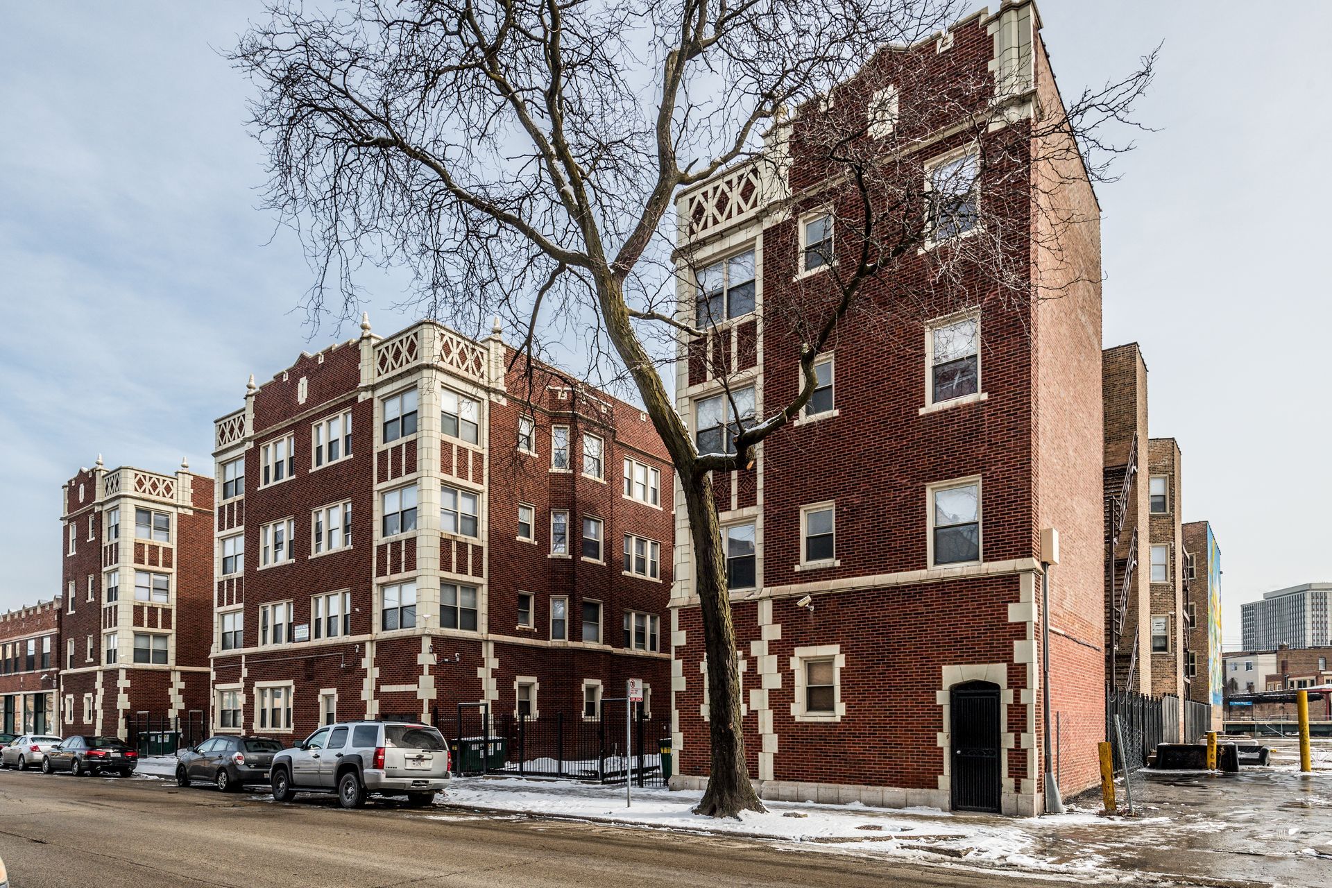 Brick apartment buildings on a snowy street with parked cars and a bare tree.