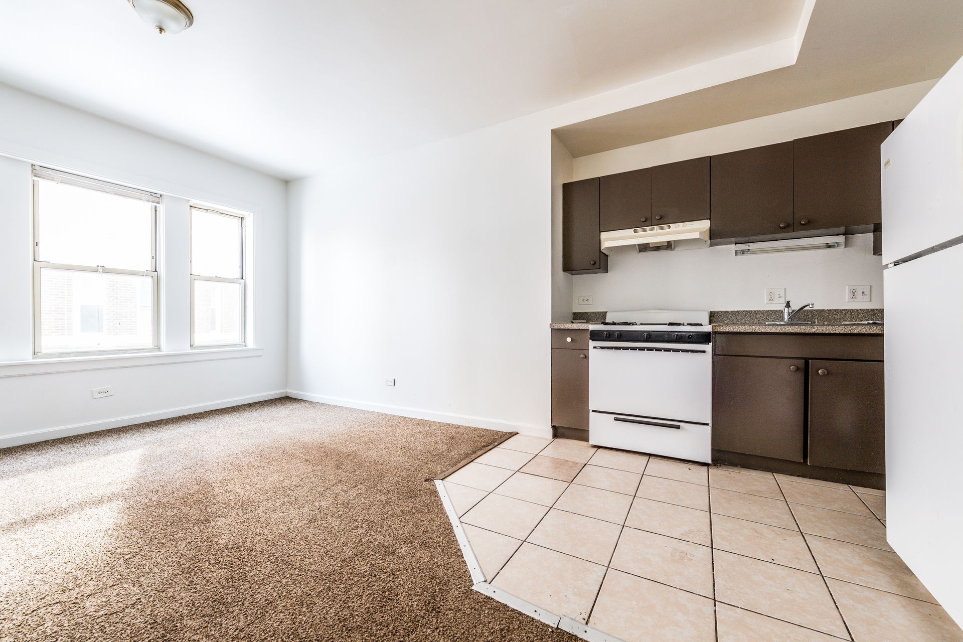Empty apartment interior with carpet and tile flooring, kitchen appliances, and windows.