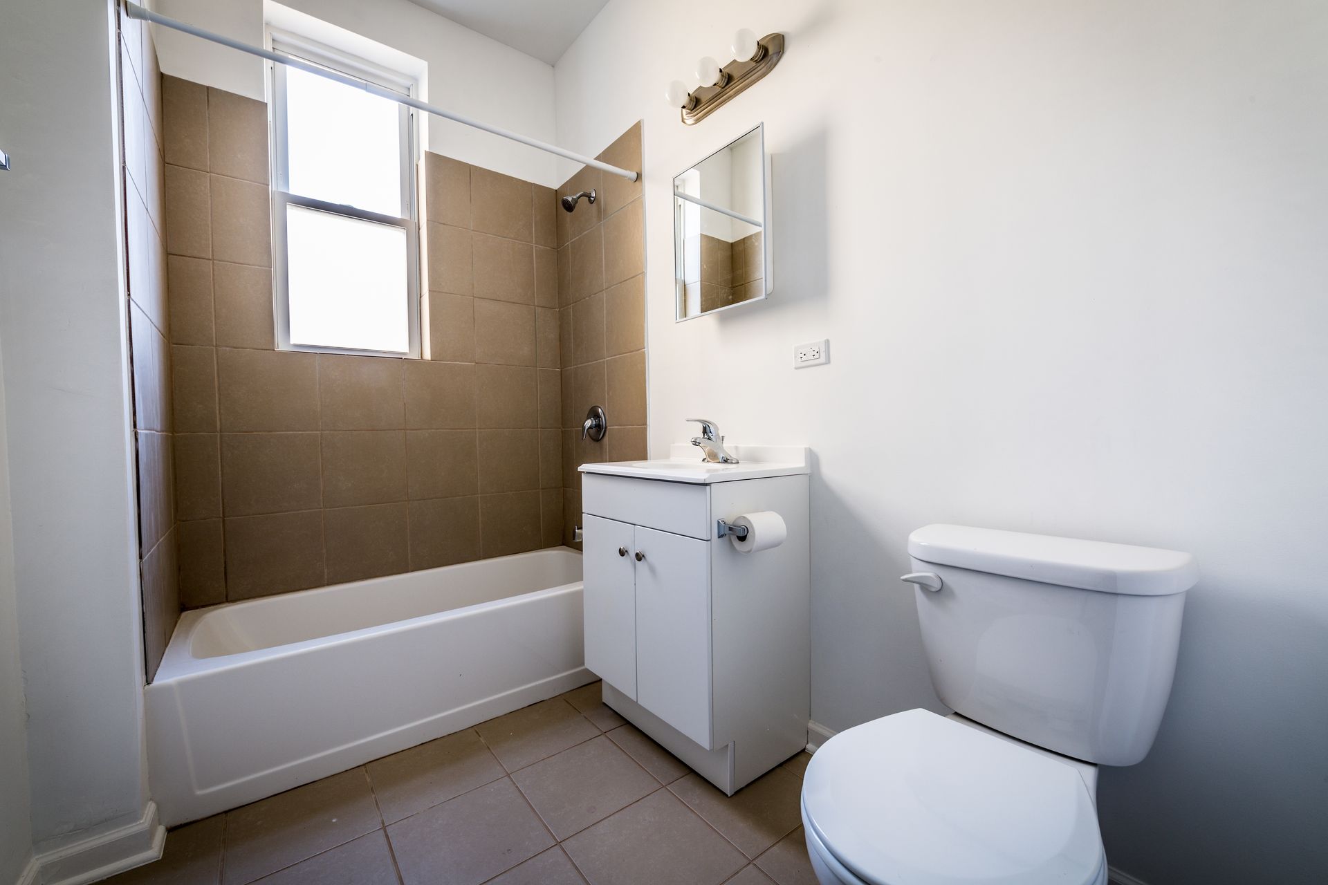 Bathroom with a white toilet, vanity, and bathtub. Brown tile in the shower area, and a window is present.