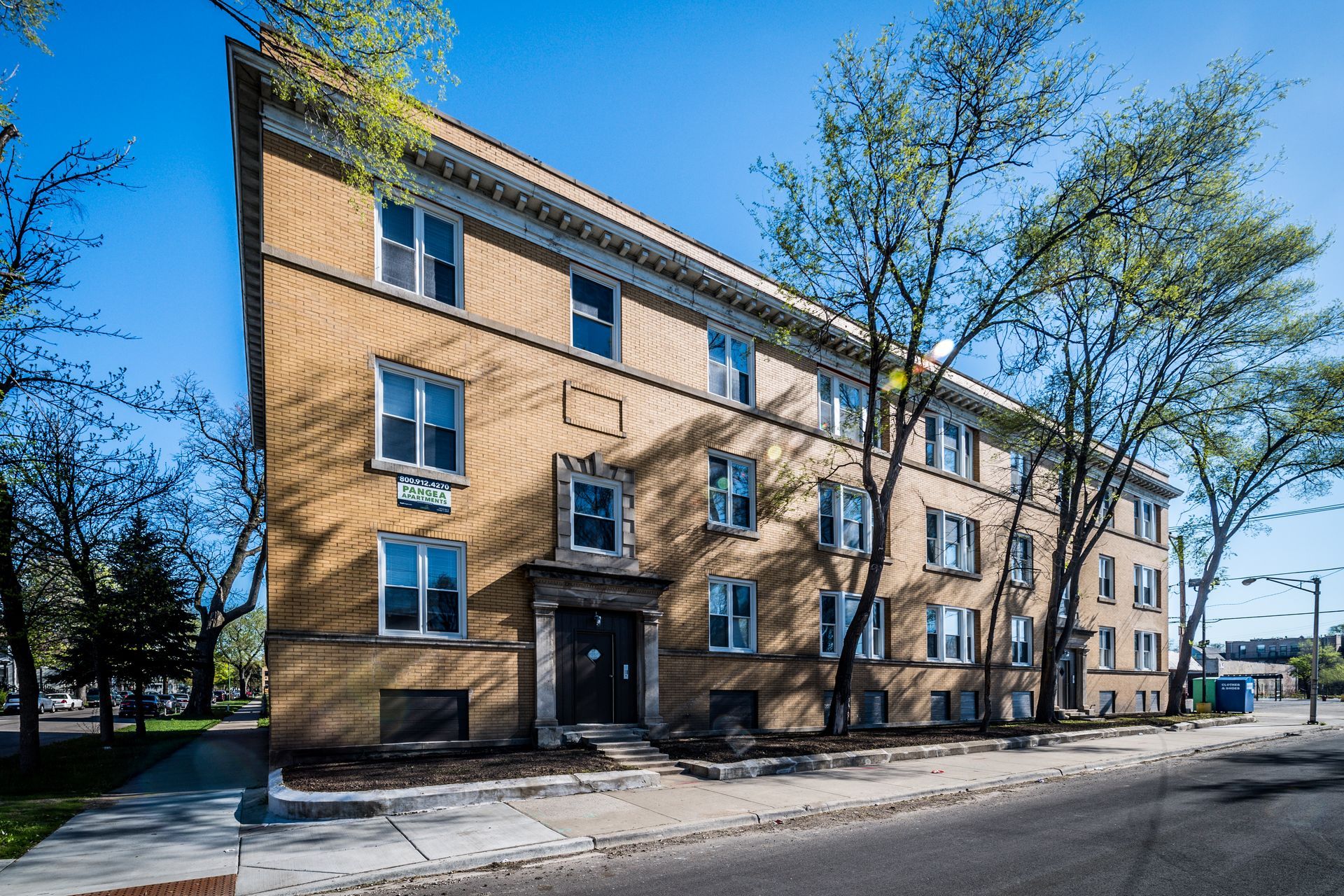 Yellow brick apartment building with multiple windows, on a sunny street, trees line the sidewalk.