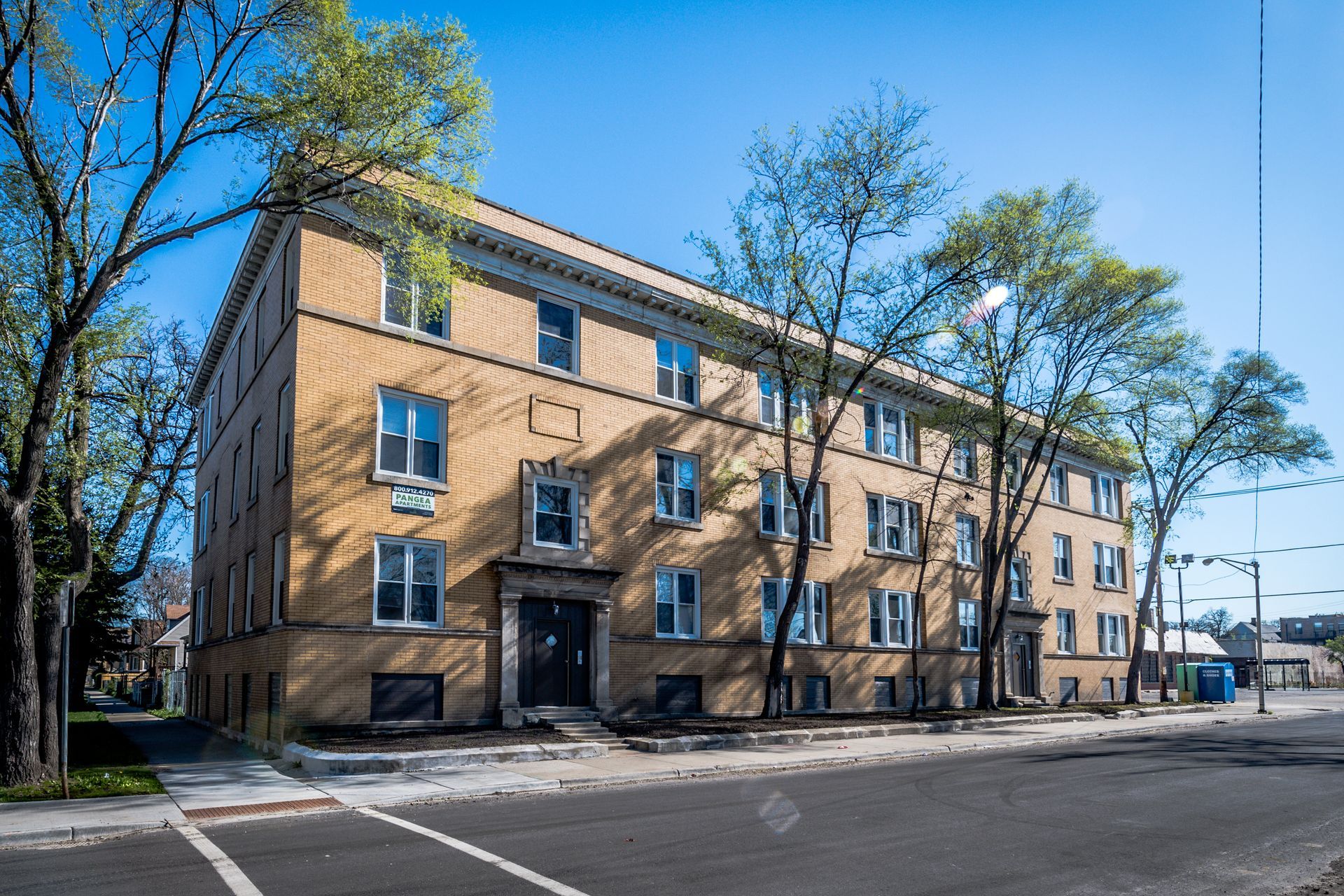 Three-story brick apartment building on a city street with trees and clear blue sky.