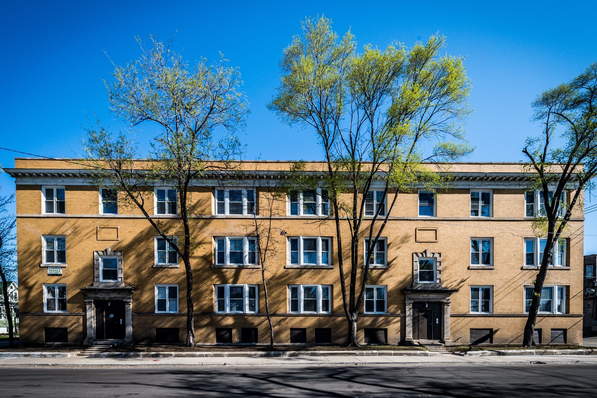 Yellow brick apartment building with white windows and trees against a blue sky.