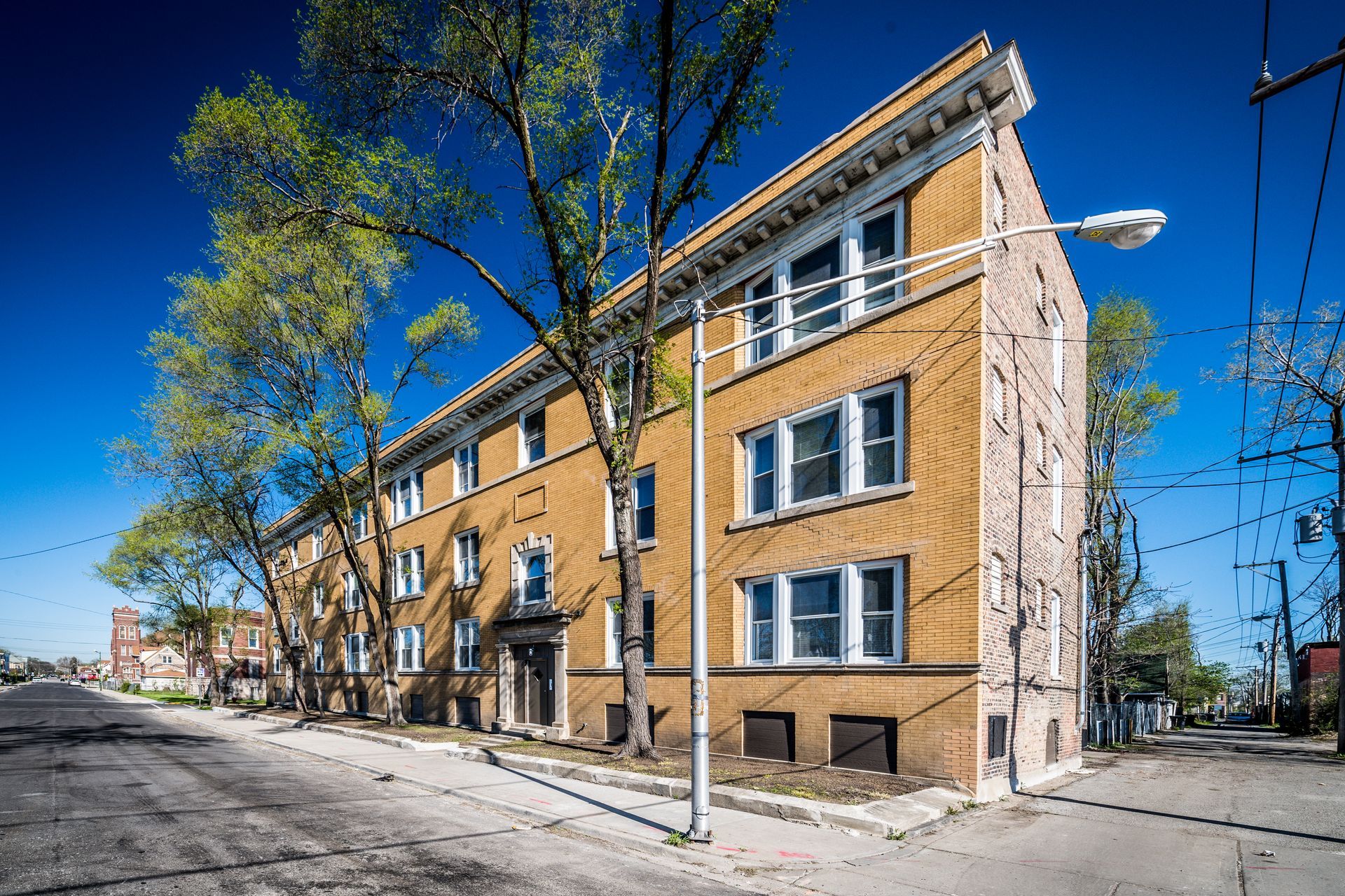 Brick apartment building on a street with a tree in front. Bright blue sky.