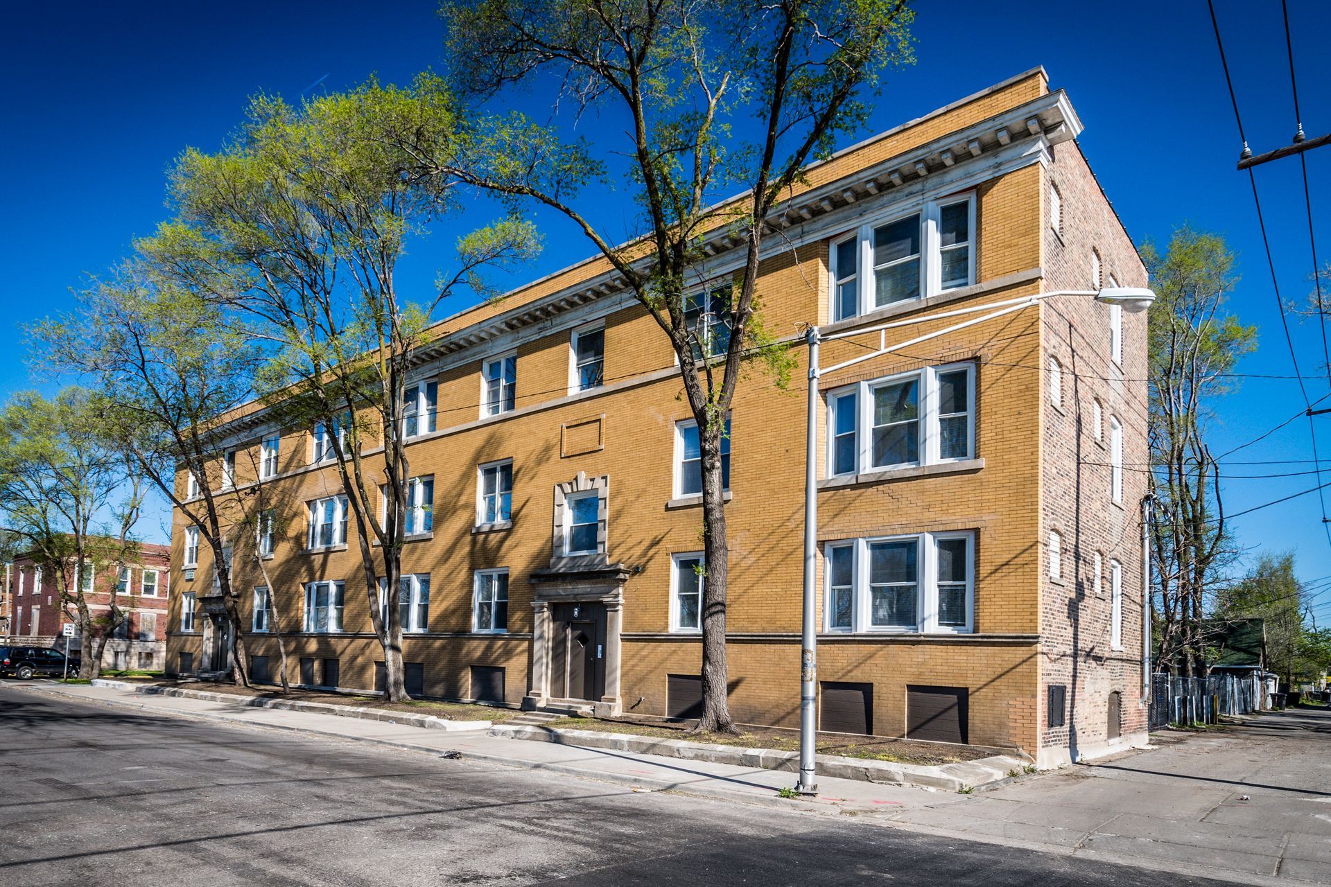 Three-story brick apartment building on a city street, trees in front, clear blue sky.