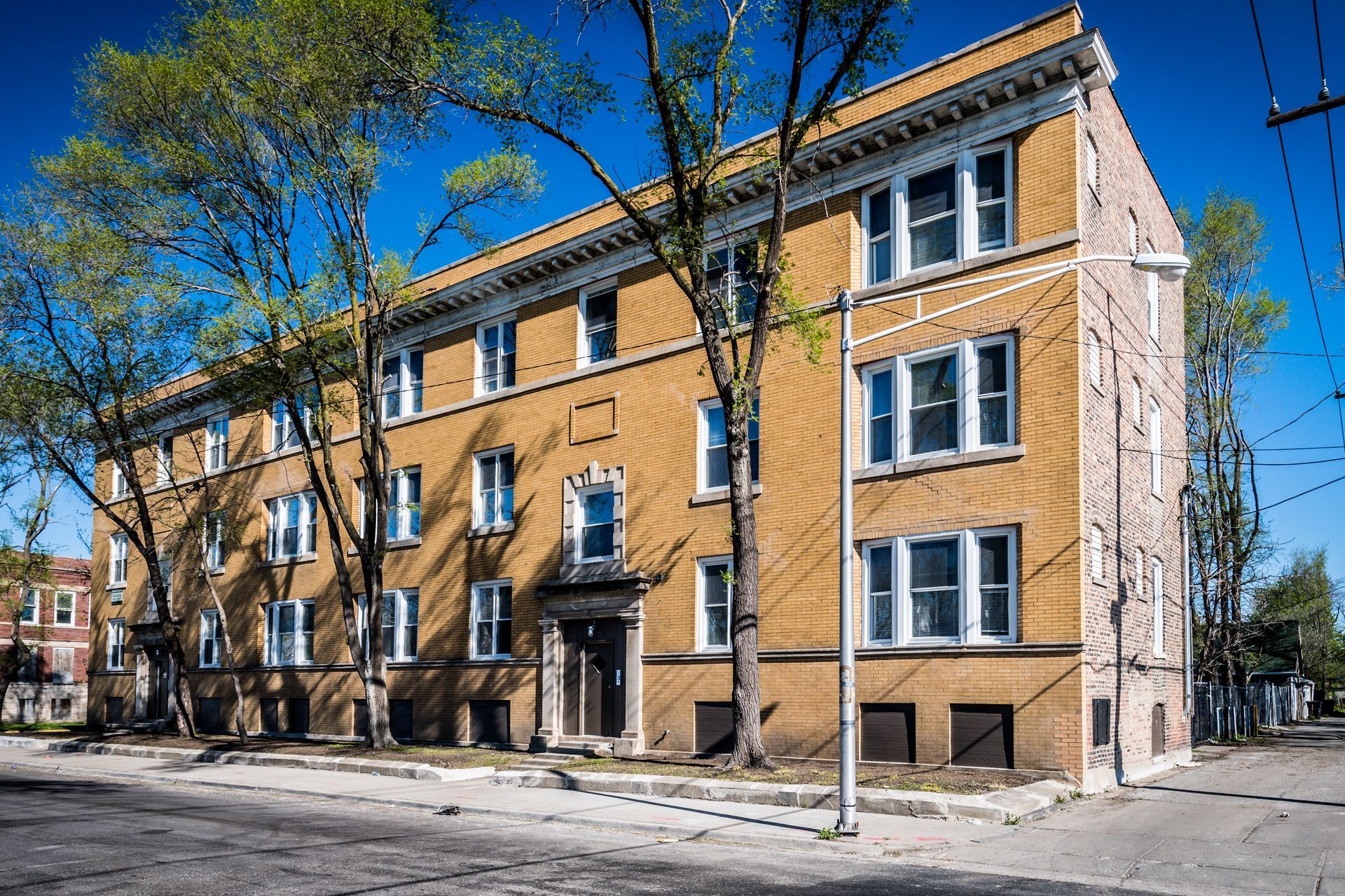 Brick apartment building on a street, with trees and a clear blue sky.