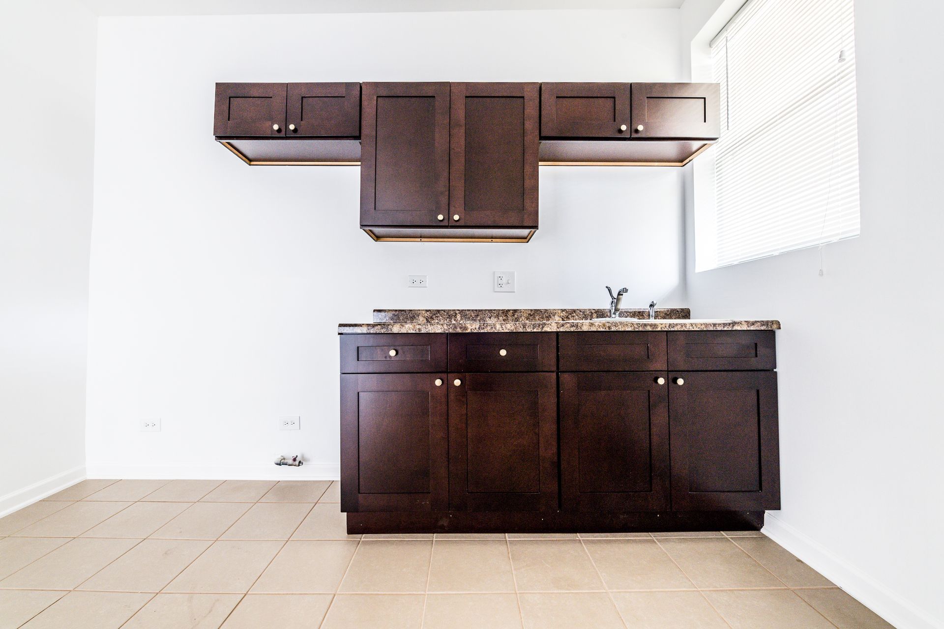 Dark brown kitchen cabinets against a white wall; granite countertop, small sink, window with blinds.