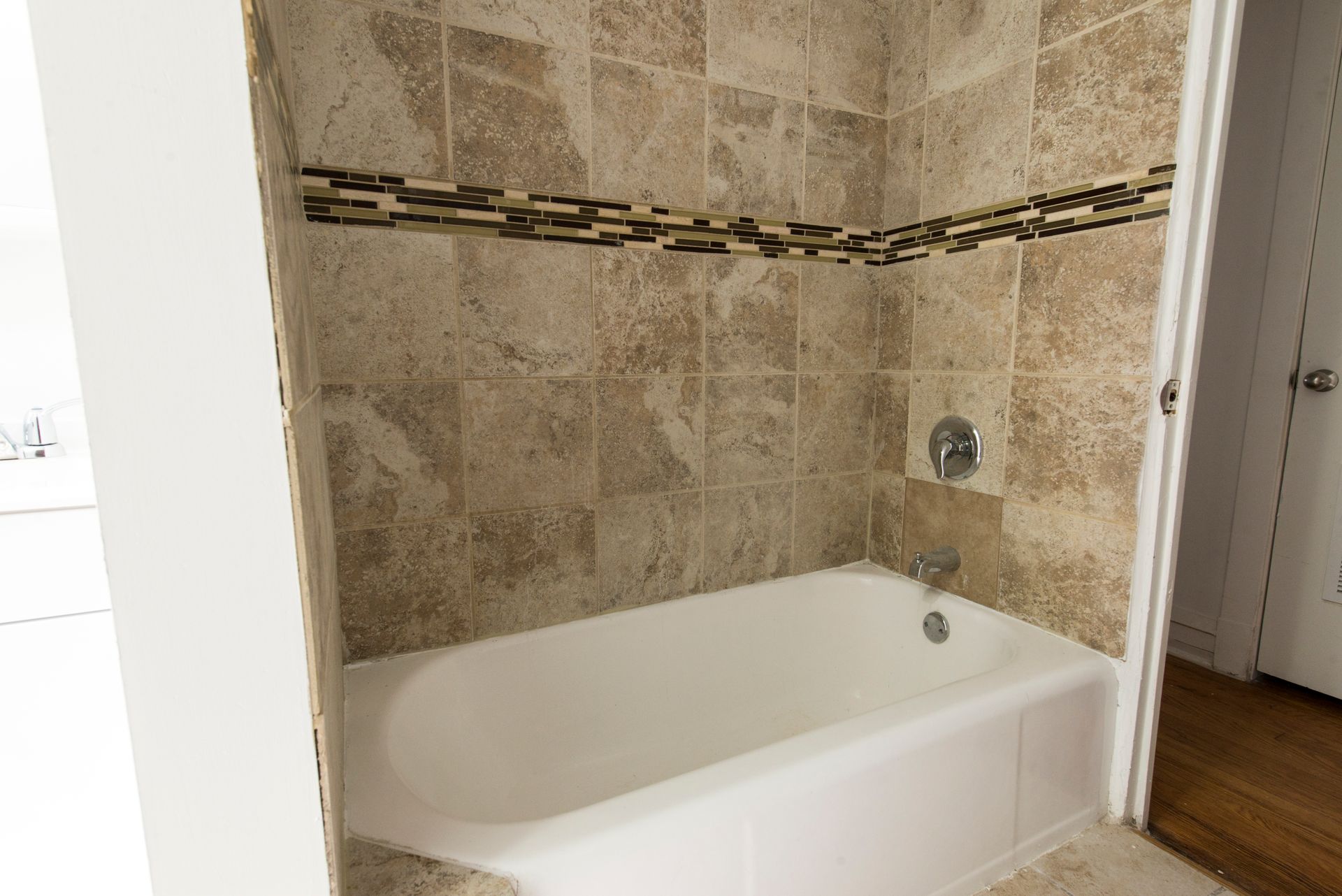 A small bathroom with a white bathtub and beige tiled walls and accent strip.