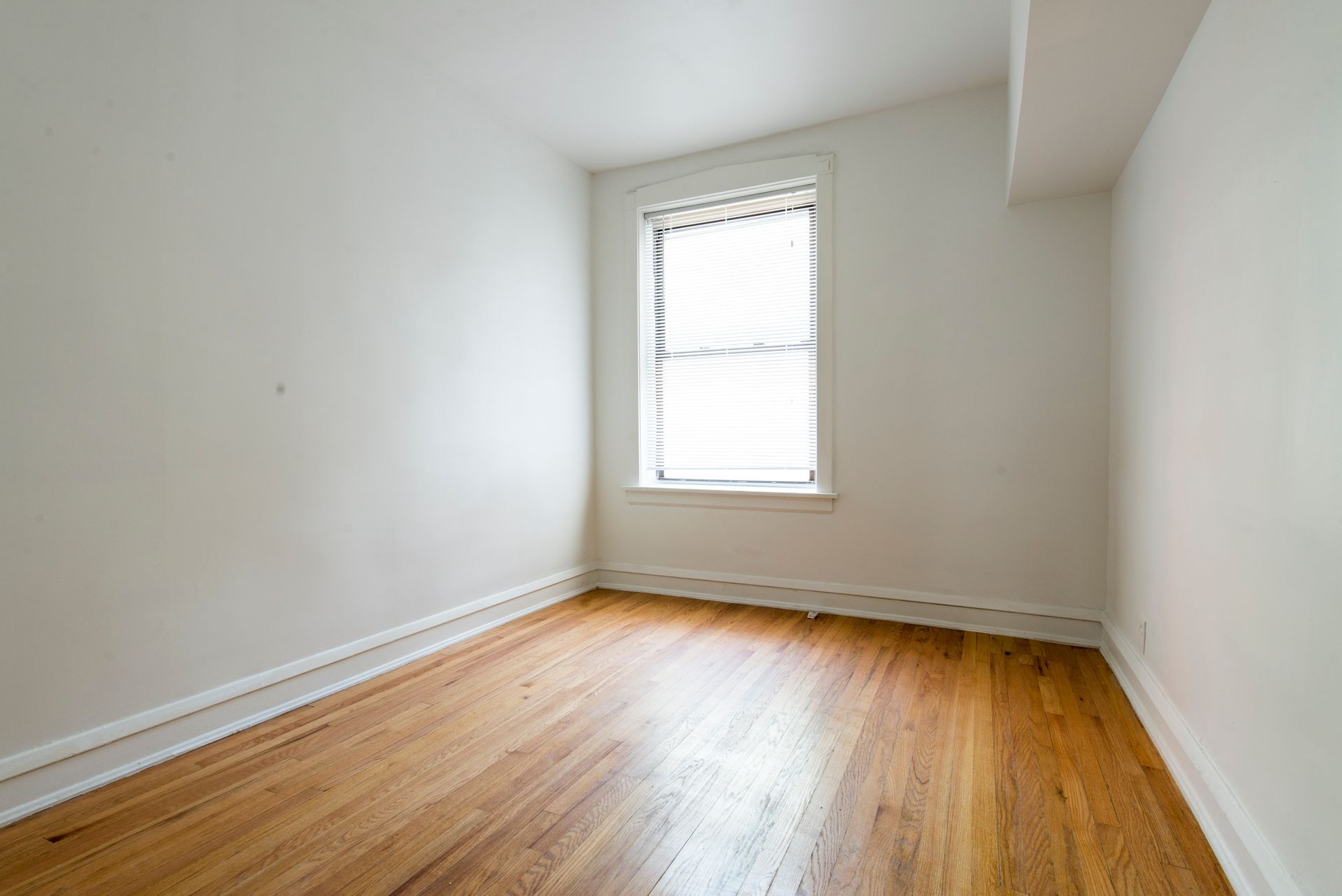 Empty room with hardwood floors, a window with blinds, and white walls.
