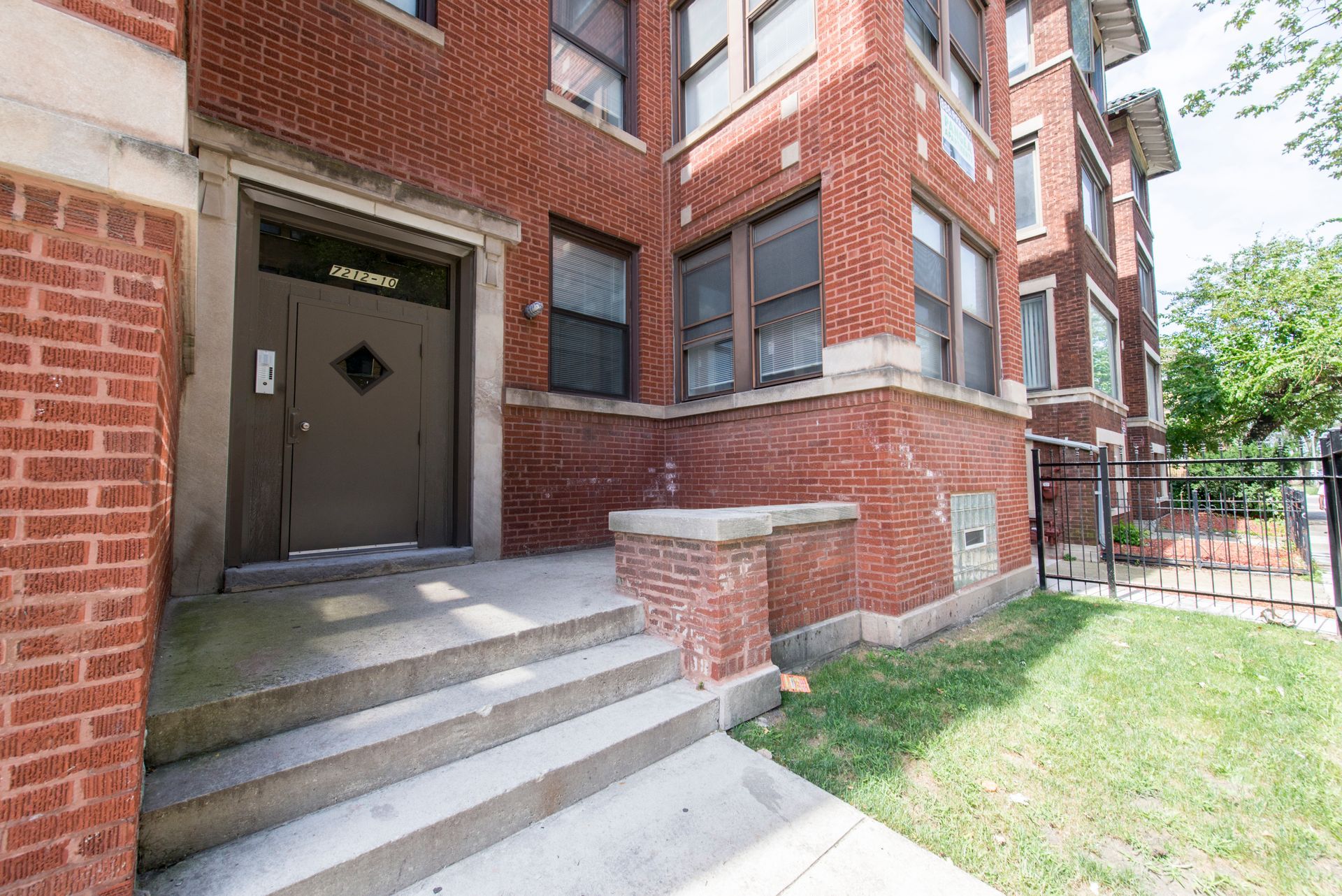 Red brick building with gray steps leading to a dark brown door, a small lawn, and a black fence.