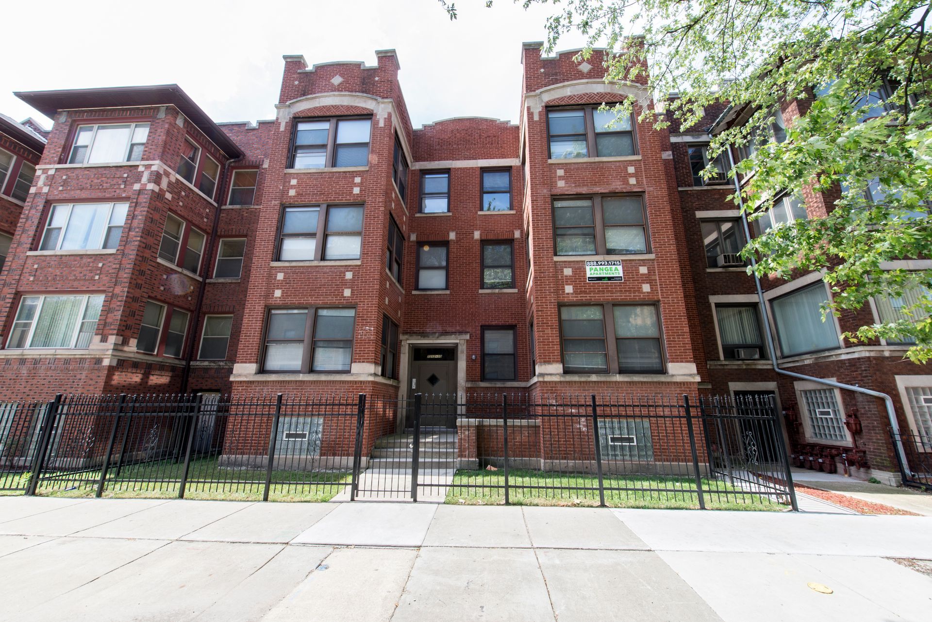 Brick apartment building with black fence, trees, and cloudy sky.