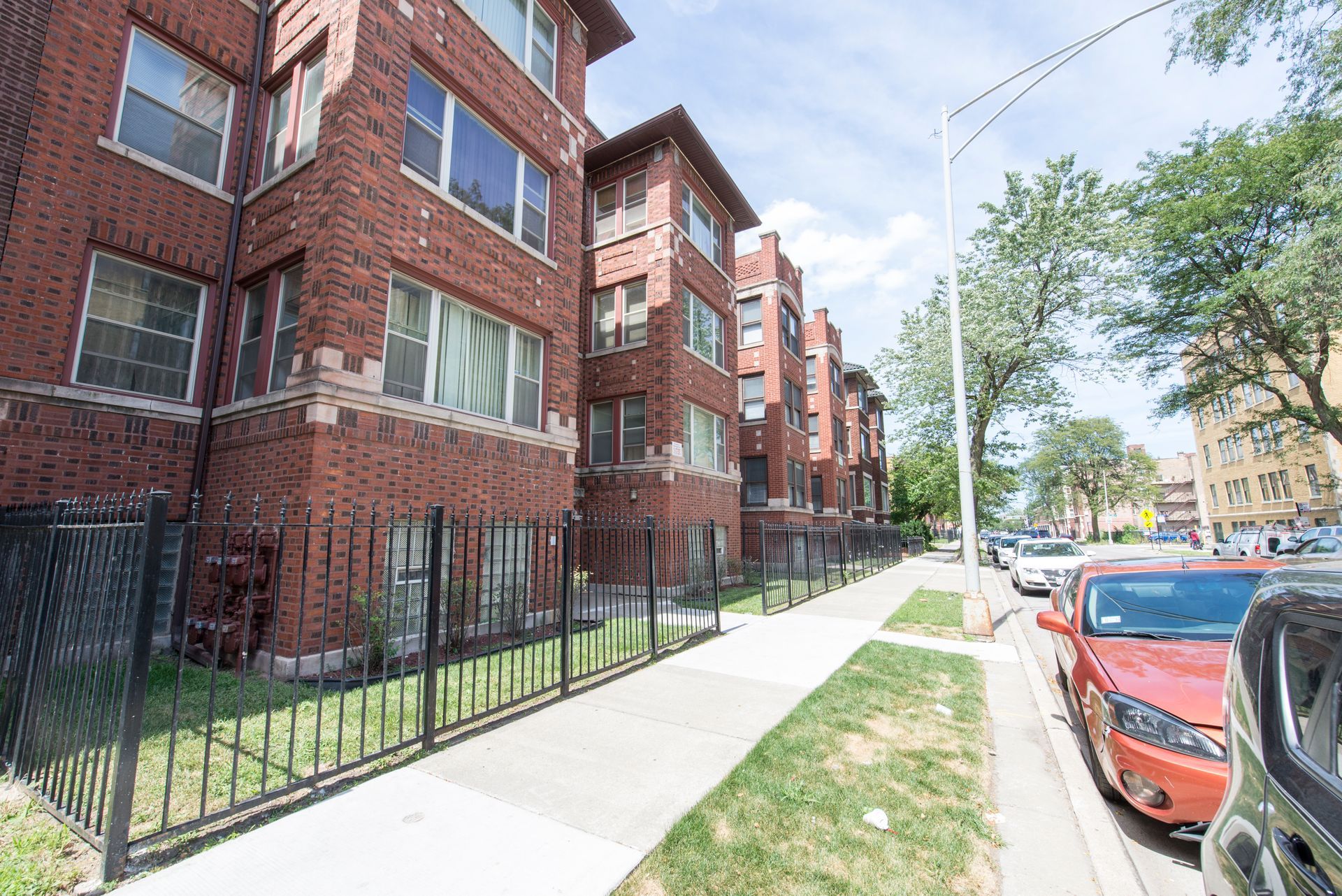 Brick apartment building with black fence, sidewalk, parked cars, and trees on a sunny street.