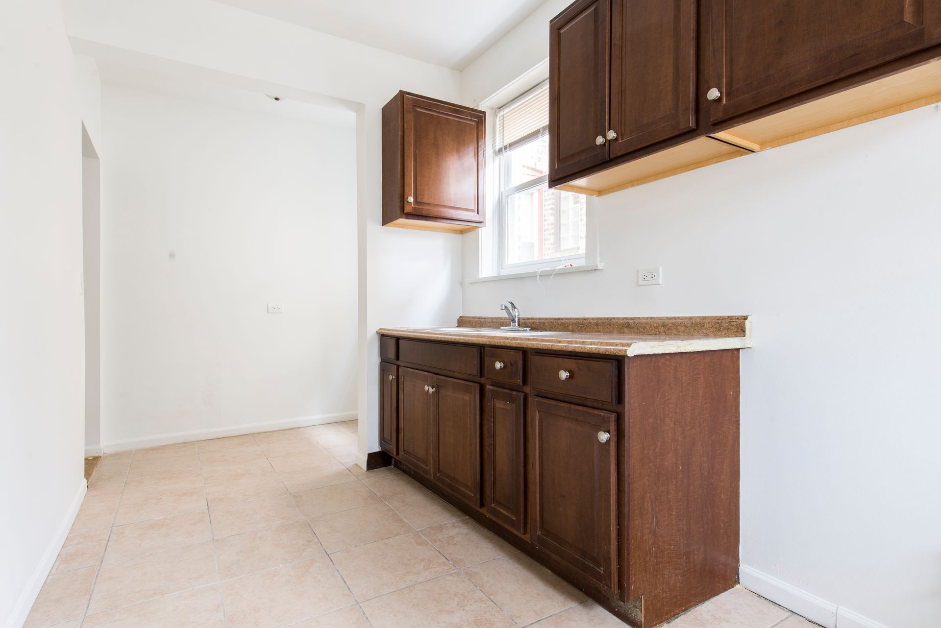Empty kitchen with brown cabinets, light countertops, and beige tiled floor. White walls.
