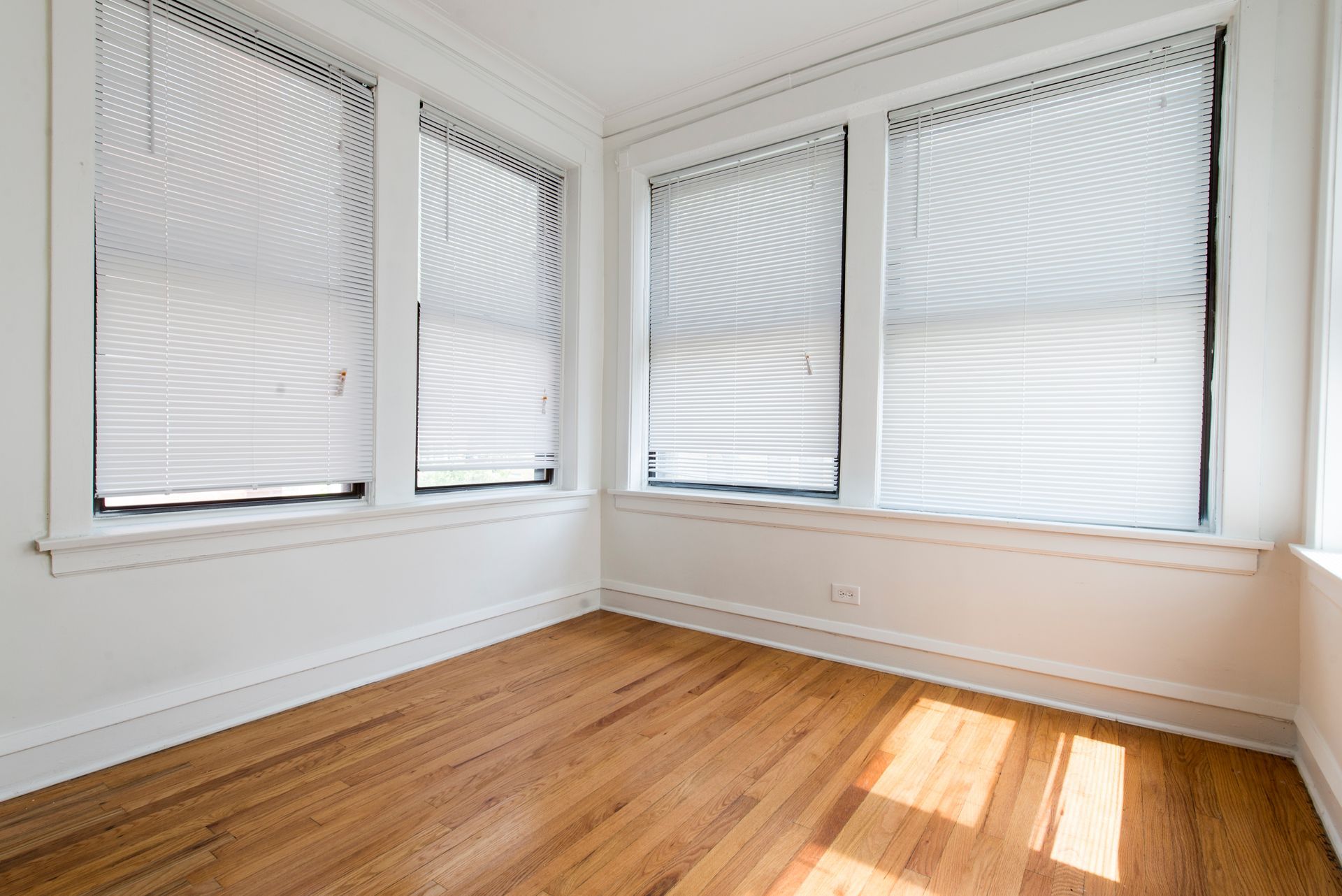 Empty room with hardwood floors, white walls, and three windows with closed blinds.