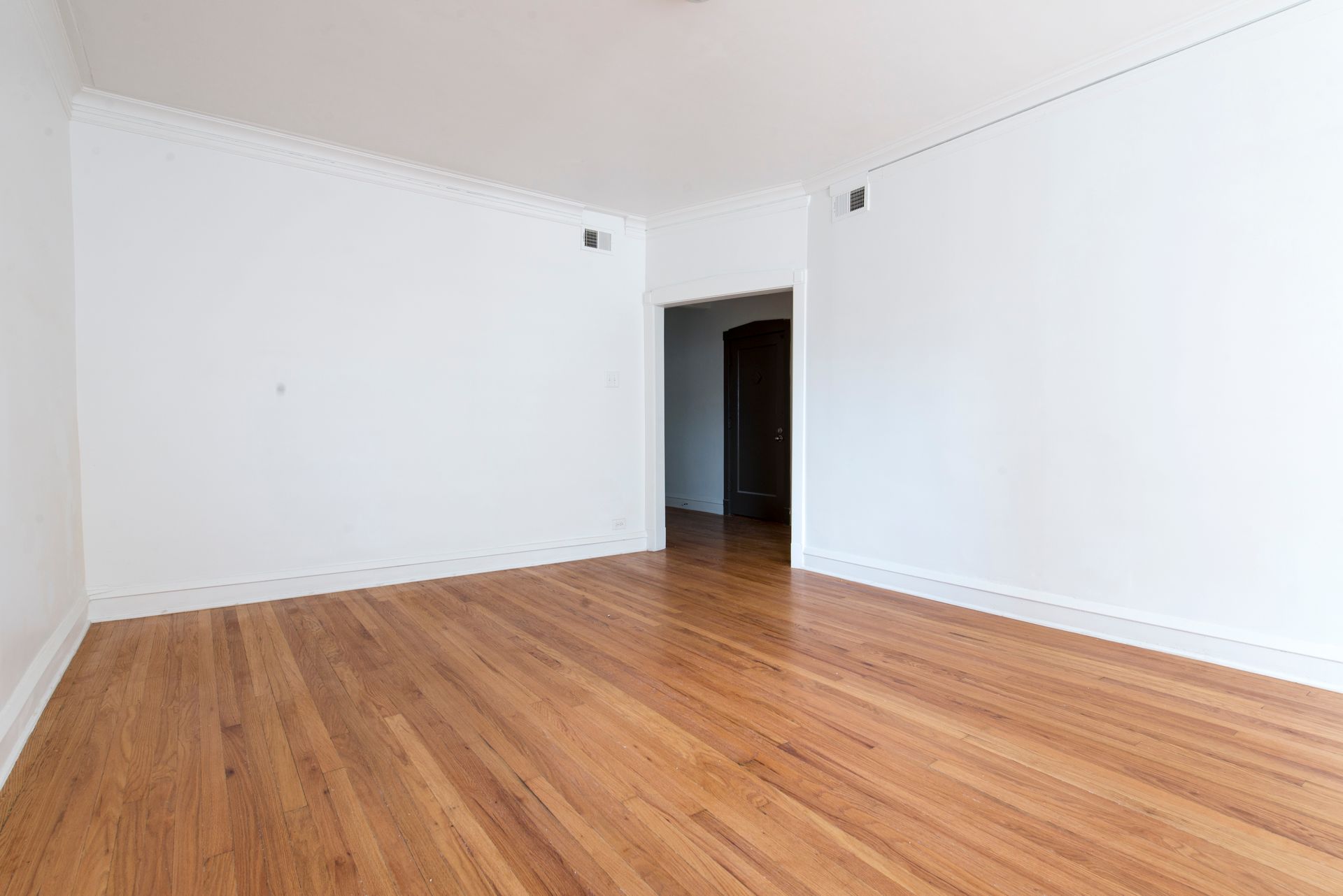 Empty room with hardwood floors, white walls, and a doorway leading to a dark hallway.