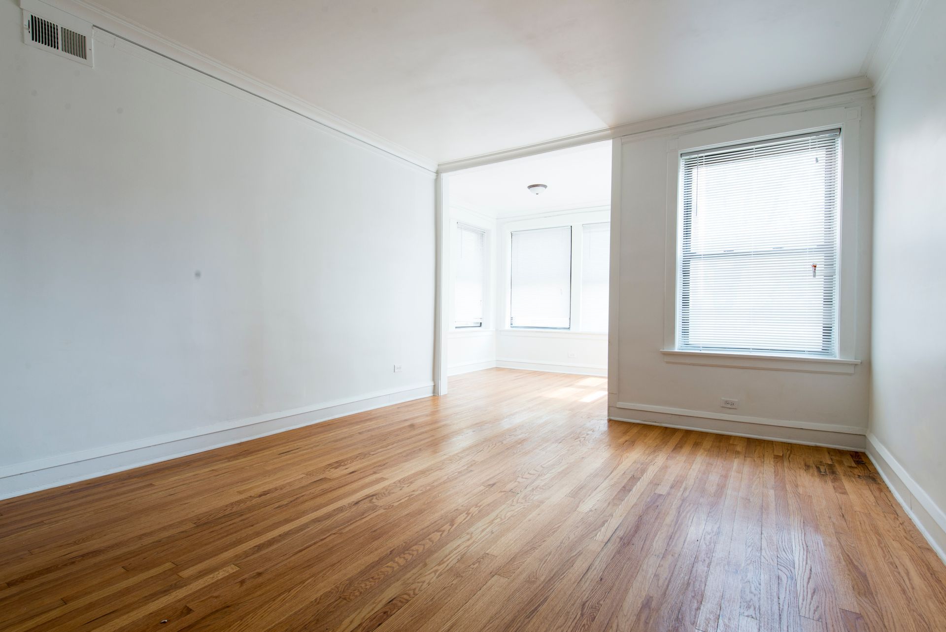 Empty room with wood floors and white walls, leading to another room with a window.