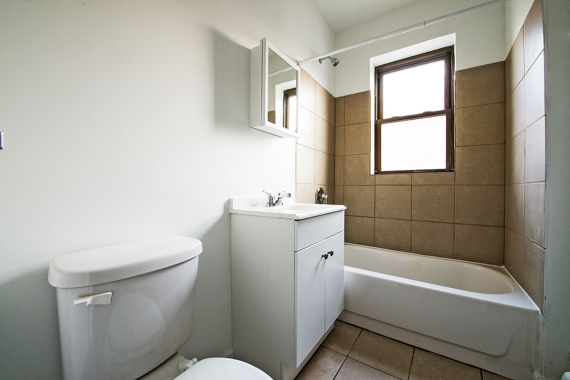 Bathroom with white toilet, vanity, and bathtub. Brown tile surrounds the window.