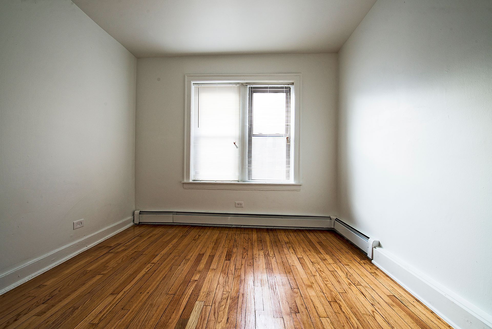 Empty room with hardwood floors, a window with blinds, and white walls.