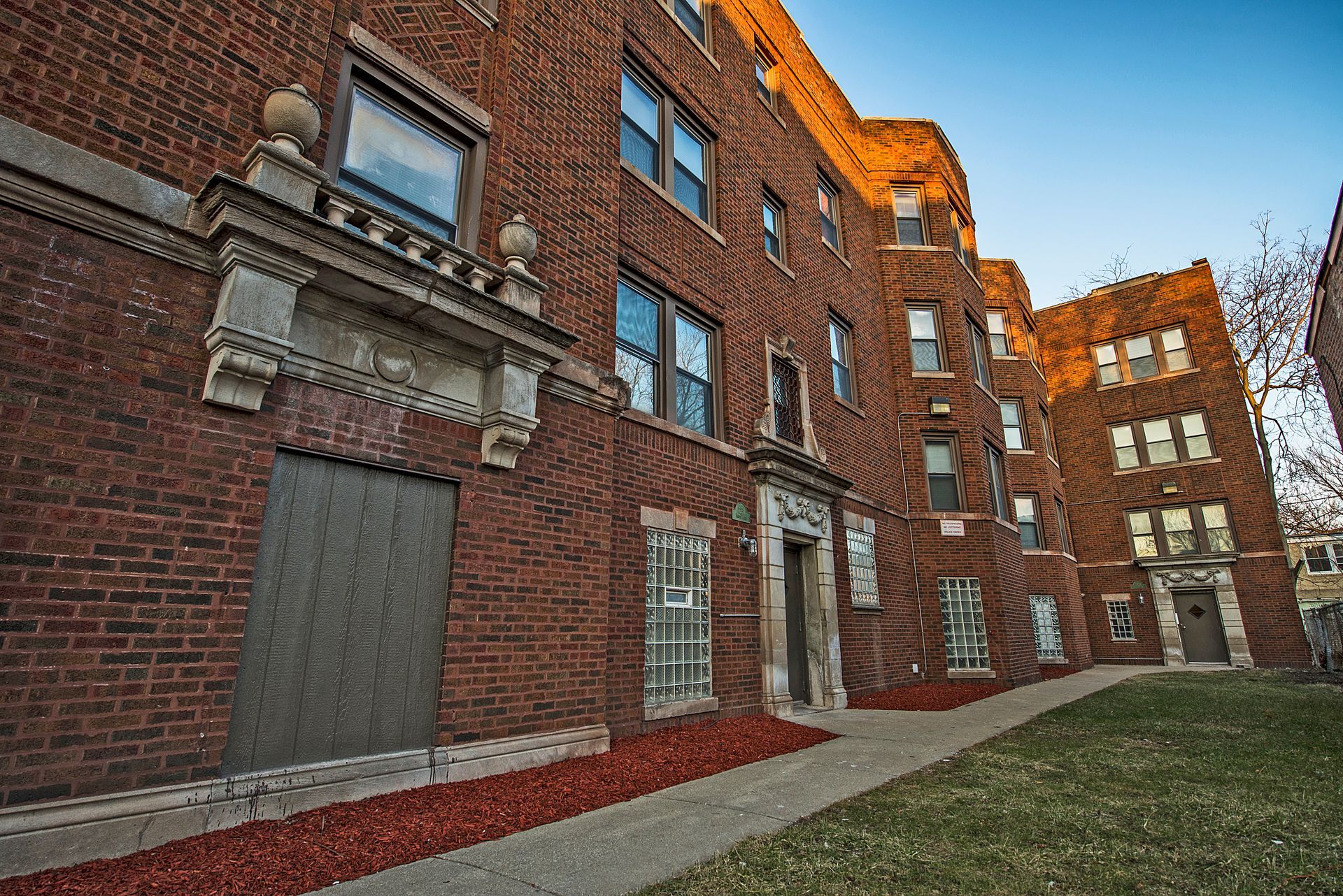 Red brick apartment building with architectural details; sidewalk and grassy area in front.