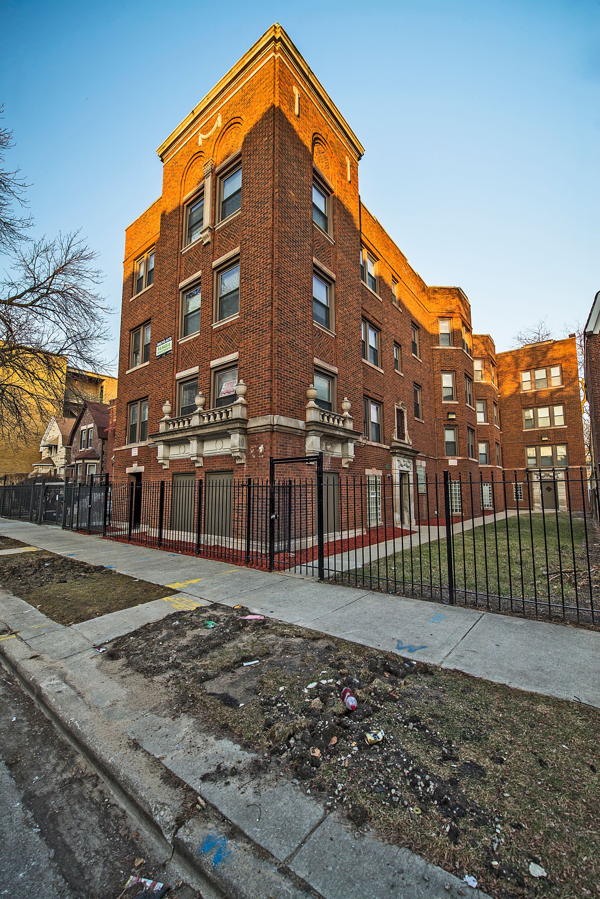 Brick apartment building, partially boarded up, behind a metal fence and sidewalk.