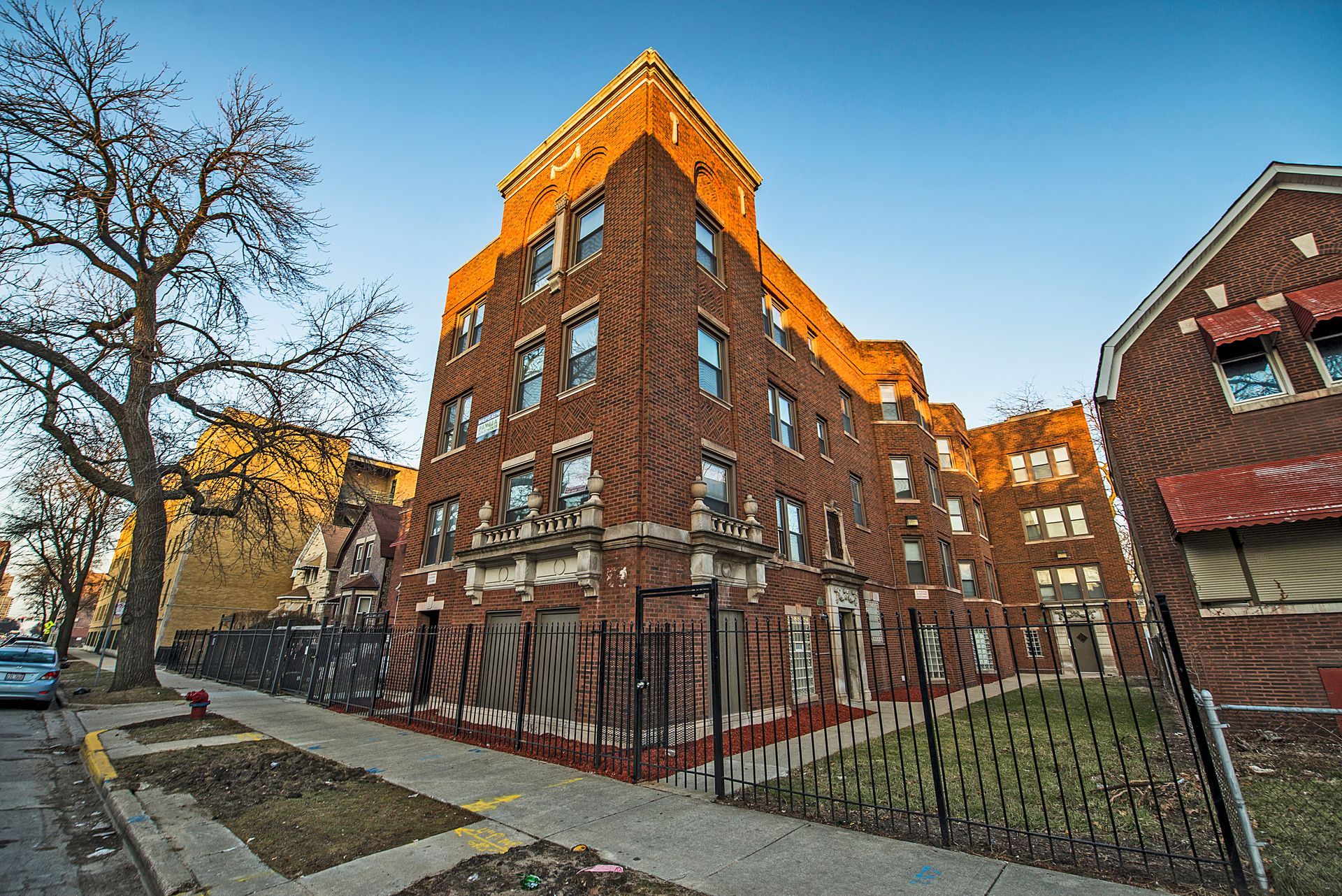 Brick apartment building with decorative stonework and wrought-iron fence on a city street.