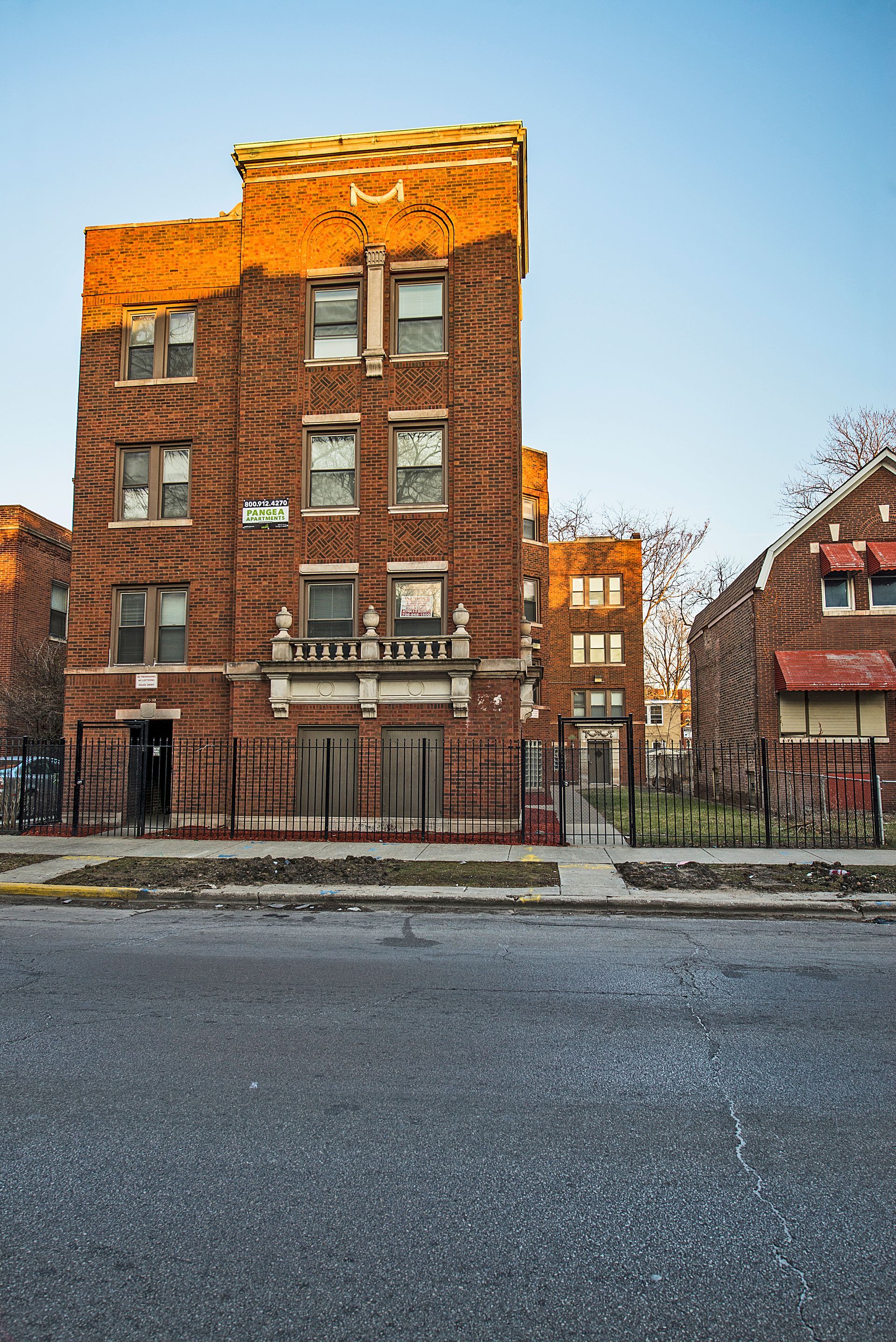 Brick apartment building in sunlight. Fenced yard, dark asphalt road in foreground.