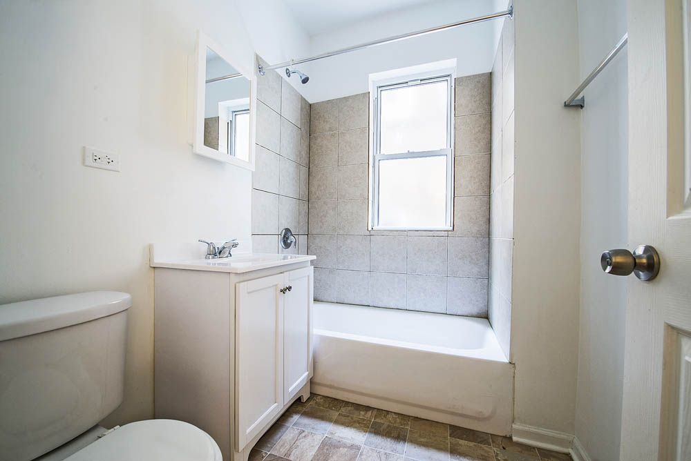 Small, white bathroom with a toilet, sink, tub, and window. Light-colored walls and floor.