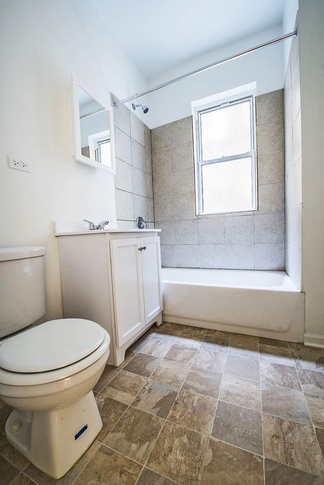 Bathroom with toilet, sink, and shower/tub combo. Light-colored walls and floor, natural light from window.