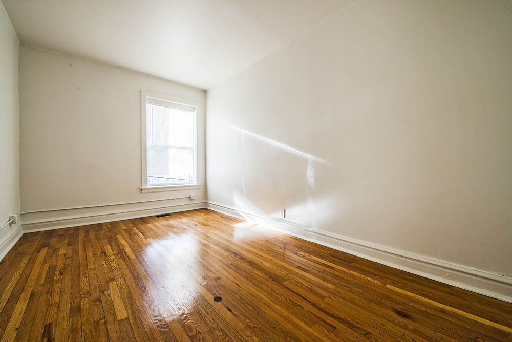 Empty room with hardwood floors, white walls, and a window. Sunlight streams across the floor.