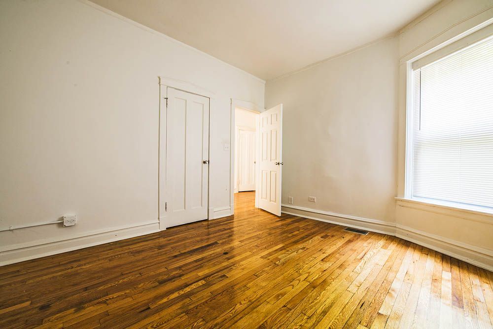 Empty room with hardwood floors, white walls, two closed doors, and a window with blinds.