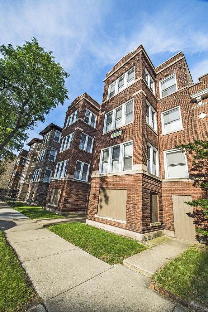 Brick apartment buildings on a city street, with sidewalk and green lawn under a blue sky.