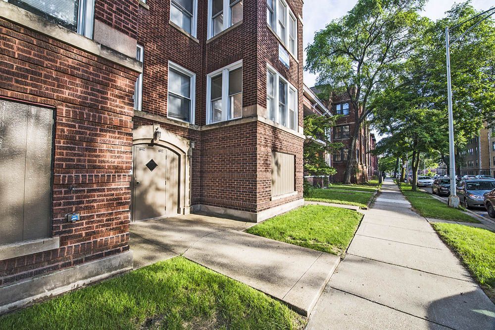 Brick apartment building with sidewalk, green grass, and street with parked cars.