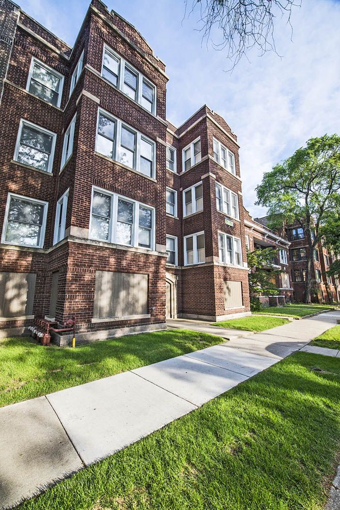 Brick apartment building with boarded-up windows, sidewalk, and green lawn under a sunny sky.