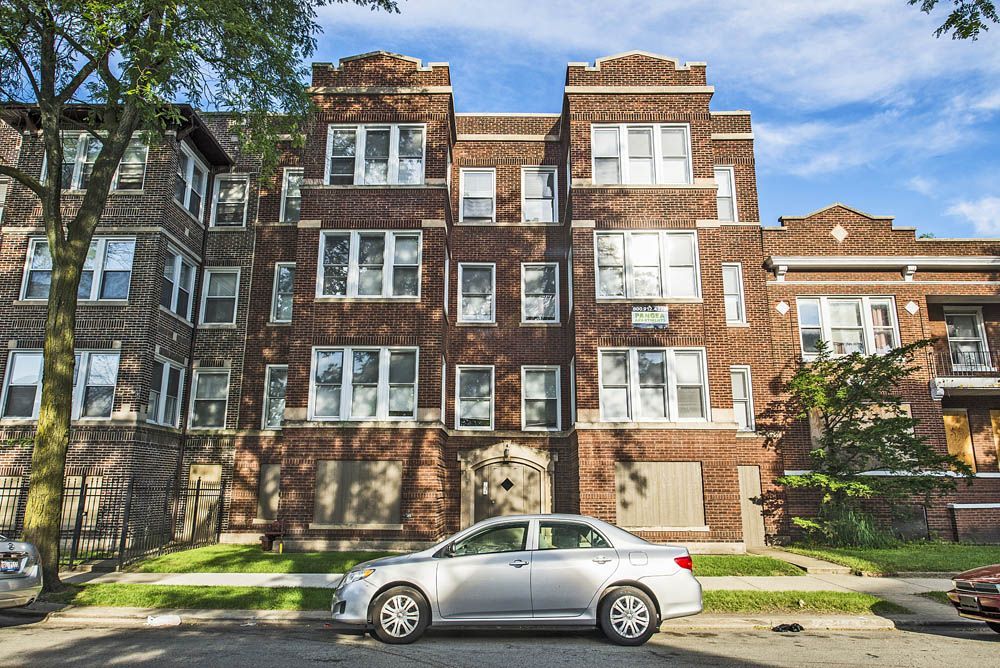 Brick apartment building with multiple windows. A silver car is parked on the street. Blue sky in the background.