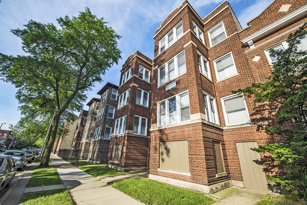 Brick apartment building on a sunny street with a green lawn and trees.