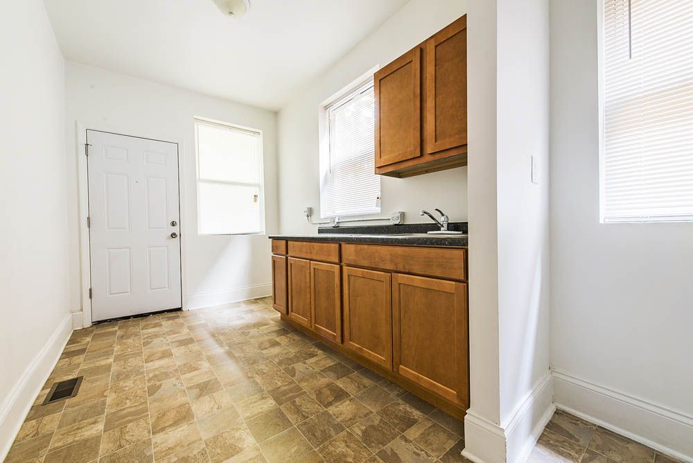 Empty kitchen with brown cabinets, a door, and two windows. Brown and tan floor. White walls.