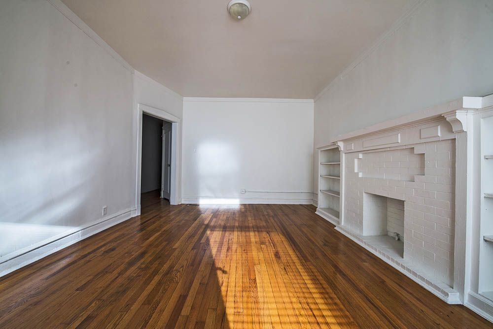 Empty room with hardwood floors, white walls, and a fireplace with bookshelves. Sunlight streams in.