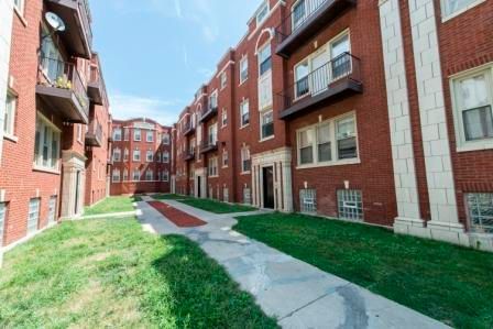 Red brick apartment building with balconies and a central walkway. Green grass and blue sky.