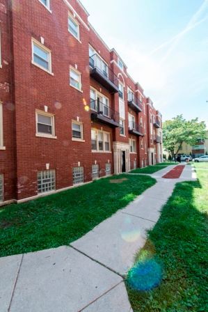 Red brick apartment building with balconies, sidewalk, and green lawn.