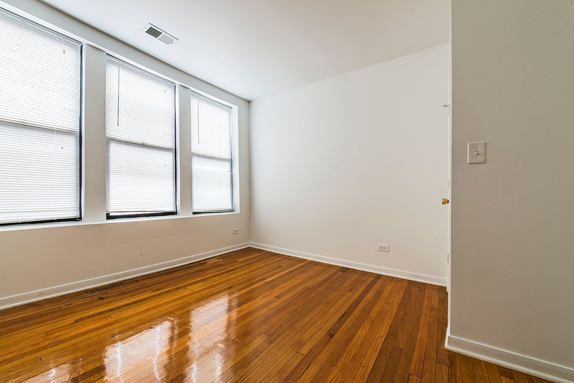 Empty room with wood flooring, three windows with blinds, and white walls.
