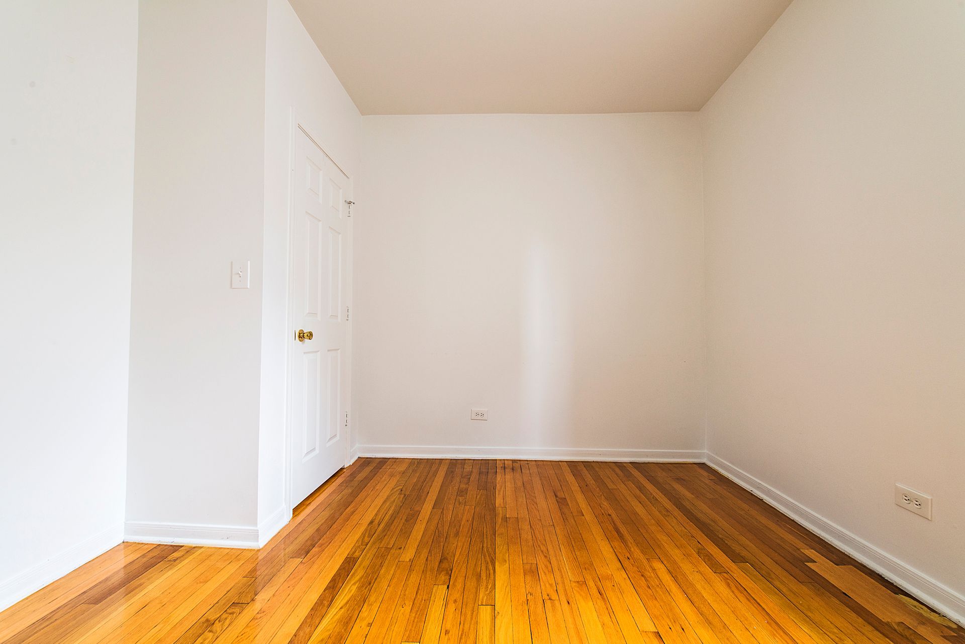 Empty room with hardwood floors and white walls. A closed door is on the left.