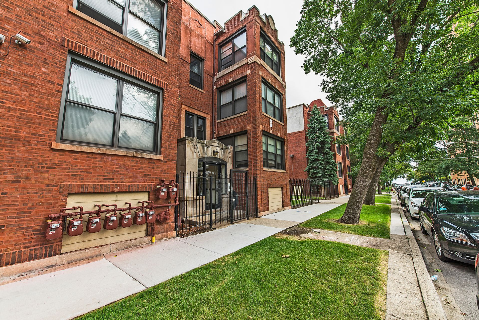 Red brick apartment building with sidewalk, green grass, and parked cars.