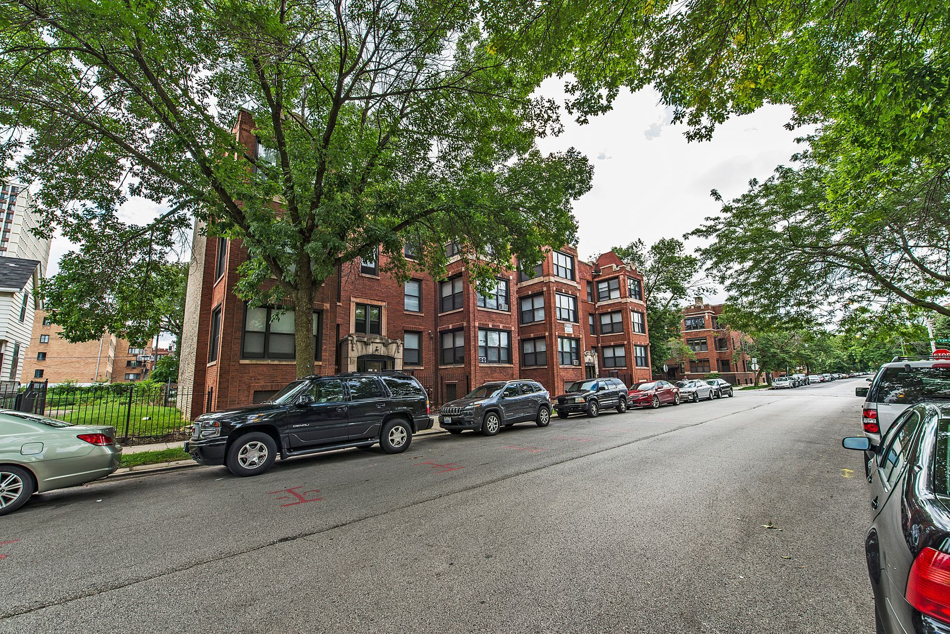Street view of brick apartment buildings with parked cars under trees.