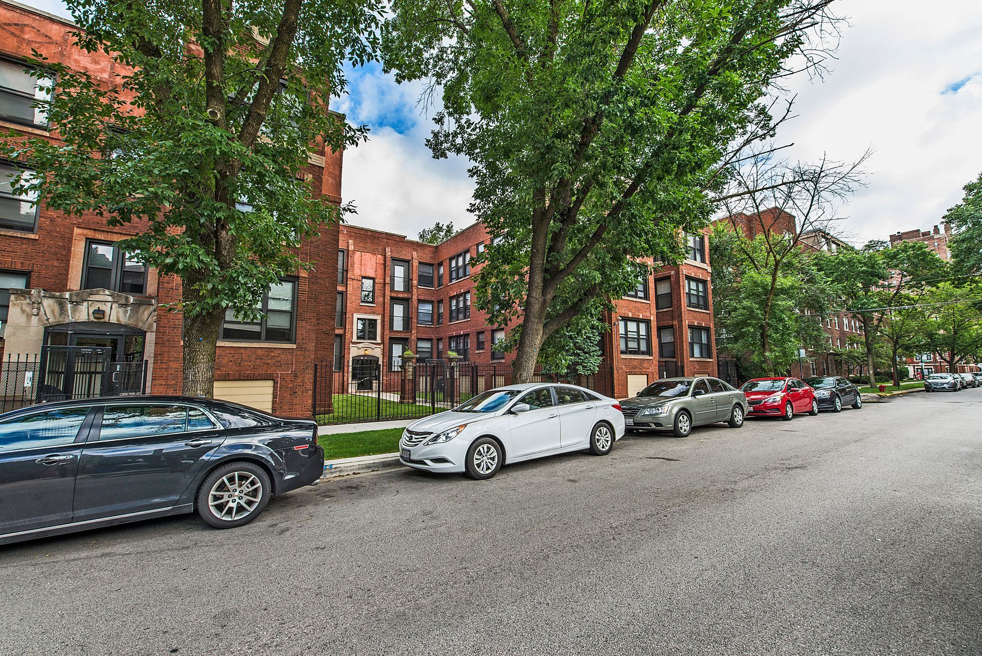 Street lined with parked cars and brick apartment buildings with green trees.
