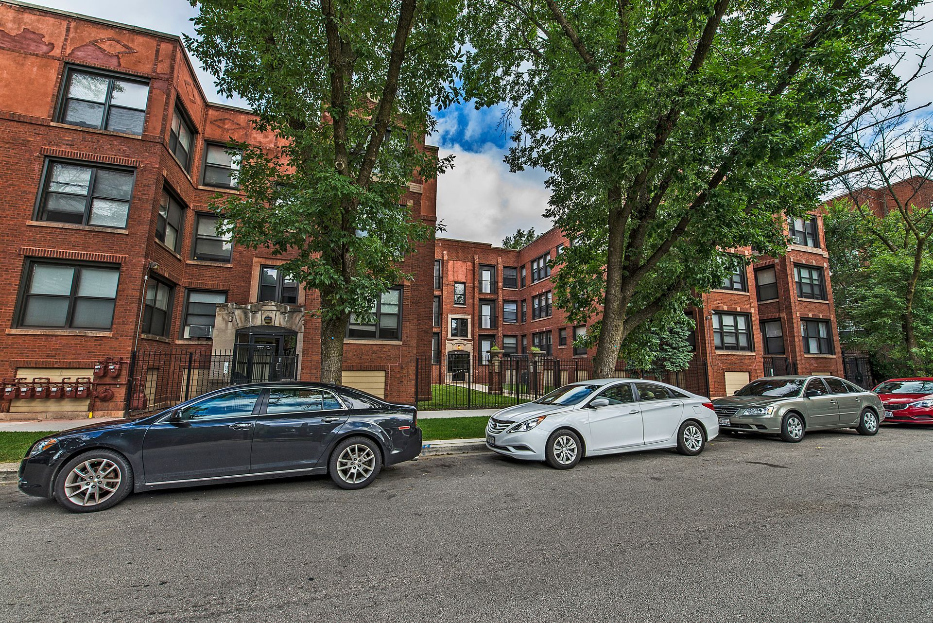 Apartment building with cars parked in front. Red brick facade, large trees, and blue sky.