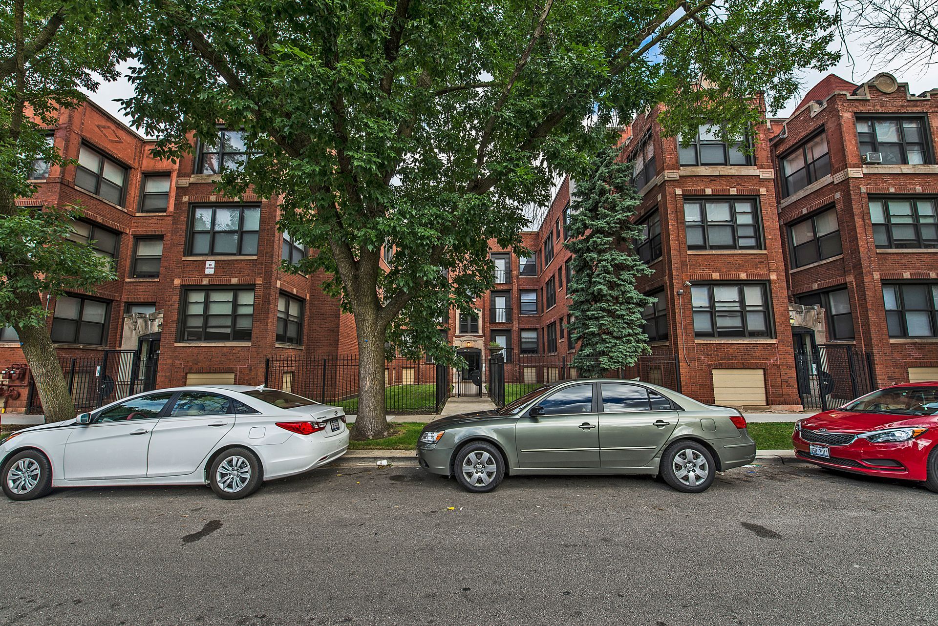 Cars parked on a street in front of brick apartment buildings with green trees.